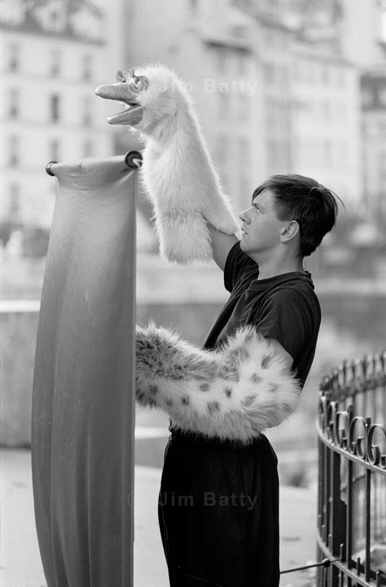 A man stands behind a tall, thin screen with a puppet on his raised right arm - in the square in front of Notre Dame Cathedral, Paris, France. He calls his act: Bululu Theatre.