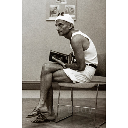 Darkly tanned man in bright white shorts, tank top and wooly hat sitting in a Parisian art gallery